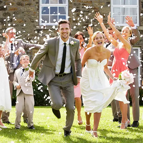 A bride and groom running through the grass with their guests.